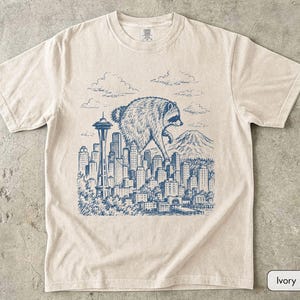 Pu&ograve; includere: Una t-shirt a maniche corte color avorio con una stampa grafica blu in stile inchiostro raffigurante un procione gigante che sovrasta lo skyline di una citt&agrave; con la Space Needle e una montagna sullo sfondo, sotto un cielo nuvoloso.