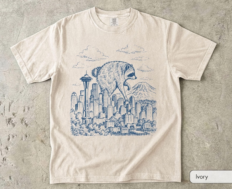 Pu&ograve; includere: Una t-shirt a maniche corte color avorio con una stampa grafica blu in stile inchiostro raffigurante un procione gigante che sovrasta lo skyline di una citt&agrave; con la Space Needle e una montagna sullo sfondo, sotto un cielo nuvoloso.