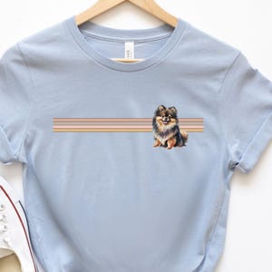Puede incluir: Camiseta azul claro con una ilustraci&oacute;n de un perro pomerania marr&oacute;n y negro y un dise&ntilde;o de rayas multicolores. El perro est&aacute; sentado y mirando hacia la derecha.
