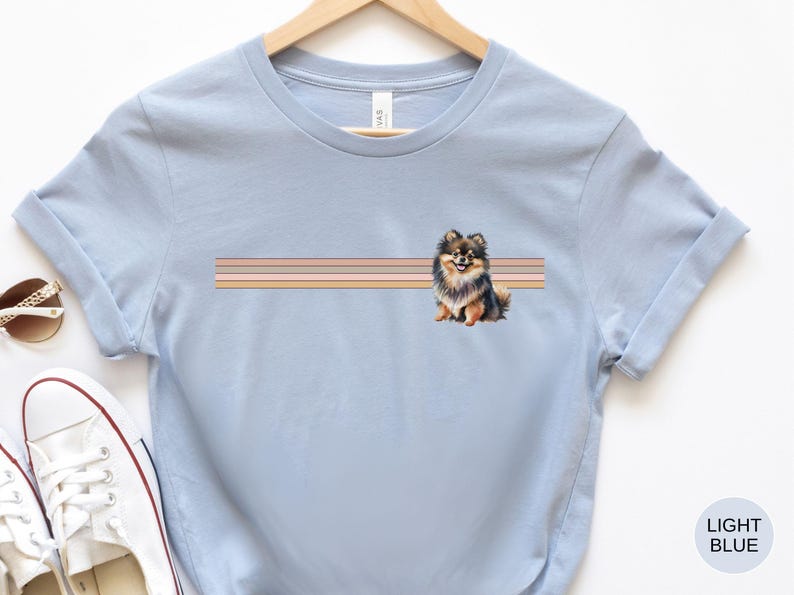 Puede incluir: Camiseta azul claro con una ilustraci&oacute;n de un perro pomerania marr&oacute;n y negro y un dise&ntilde;o de rayas multicolores. El perro est&aacute; sentado y mirando hacia la derecha.