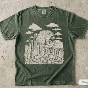 Pu&ograve; includere: Una t-shirt a maniche corte con girocollo in un colore verde oliva tenue. La parte anteriore presenta un'illustrazione grafica color crema di un procione gigante che sovrasta lo skyline di una citt&agrave;, inclusi lo Space Needle e una montagna.