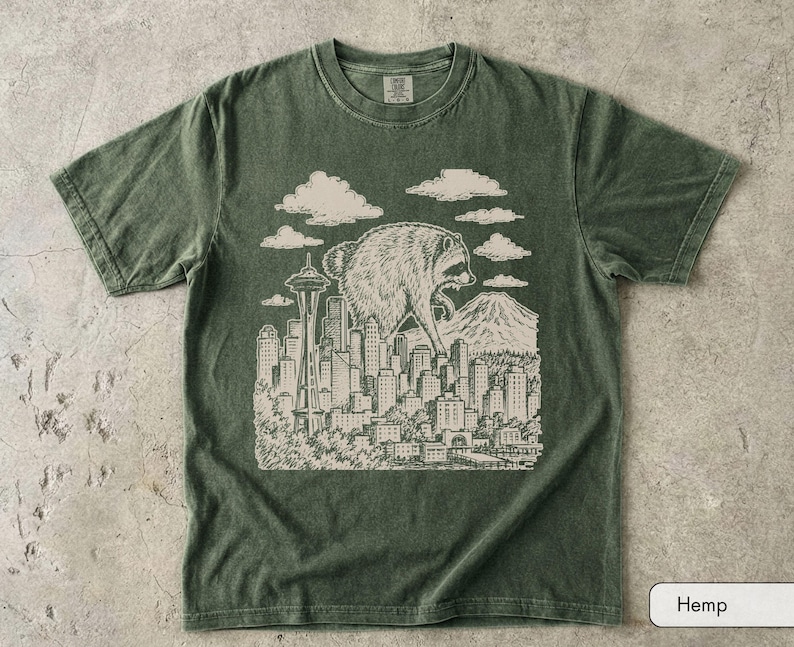 Pu&ograve; includere: Una t-shirt a maniche corte con girocollo in un colore verde oliva tenue. La parte anteriore presenta un'illustrazione grafica color crema di un procione gigante che sovrasta lo skyline di una citt&agrave;, inclusi lo Space Needle e una montagna.