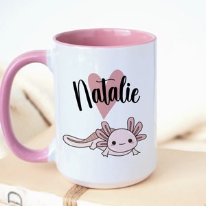 K&ouml;nnte beinhalten: Wei&szlig;er Keramikbecher mit rosa Innenraum und Griff. Der Becher zeigt ein rosa Herz mit dem Namen "Natalie" in schwarzer Schrift und eine Cartoon-Illustration eines Axolotl. Der Becher steht auf einem Stapel B&uuml;cher.