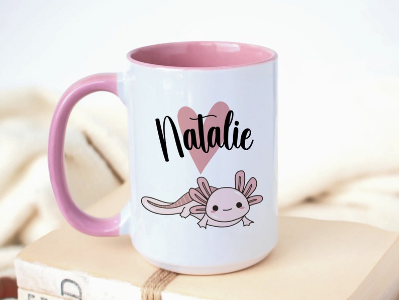 K&ouml;nnte beinhalten: Wei&szlig;er Keramikbecher mit rosa Innenraum und Griff. Der Becher zeigt ein rosa Herz mit dem Namen "Natalie" in schwarzer Schrift und eine Cartoon-Illustration eines Axolotl. Der Becher steht auf einem Stapel B&uuml;cher.
