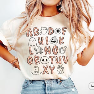 May include: An ivory t-shirt featuring a graphic print of the alphabet with letters in a red and white checkered pattern. The design includes Halloween-themed illustrations such as a ghost, pumpkin, mummy, potion bottle, skull, hat, and hand.