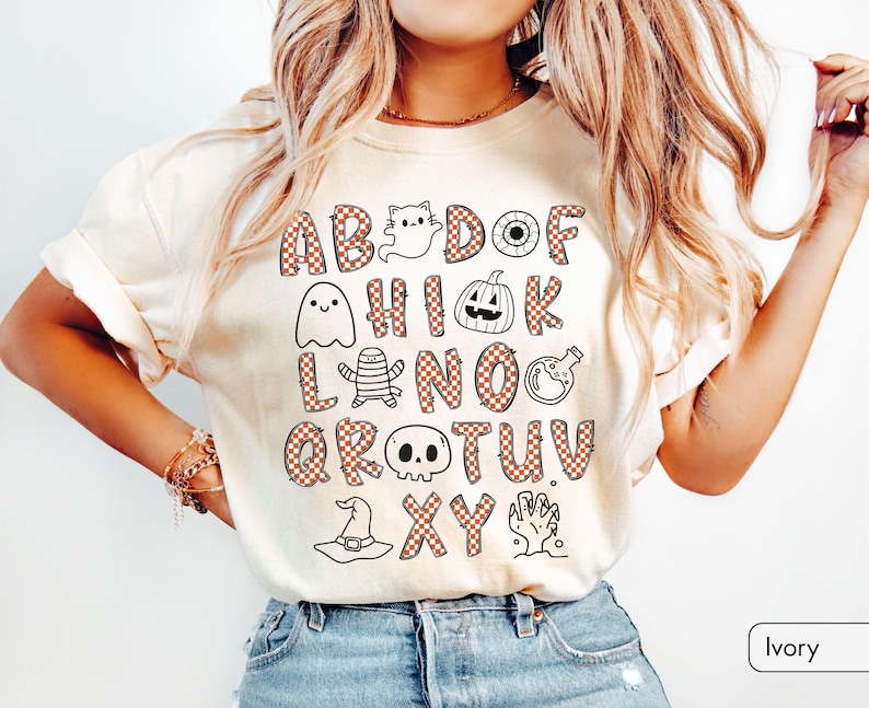 May include: An ivory t-shirt featuring a graphic print of the alphabet with letters in a red and white checkered pattern. The design includes Halloween-themed illustrations such as a ghost, pumpkin, mummy, potion bottle, skull, hat, and hand.