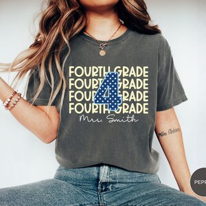 May include: A charcoal gray short-sleeve t-shirt featuring the text FOURTH GRADE repeated four times in light yellow. A large blue number 4 with white polka dots is centered over the text, with the script text Mrs. Smith printed below.