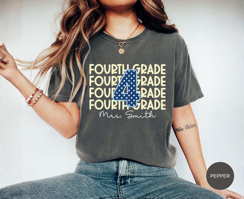 May include: A charcoal gray short-sleeve t-shirt featuring the text FOURTH GRADE repeated four times in light yellow. A large blue number 4 with white polka dots is centered over the text, with the script text Mrs. Smith printed below.