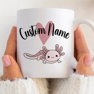 K&ouml;nnte beinhalten: Wei&szlig;e Keramik-Tasse mit einer rosa Axolotl-Illustration und einem rosa Herz. Der Text "Custom Name" ist in schwarzer Schrift geschrieben. Die Tasse hat einen wei&szlig;en Henkel, ideal f&uuml;r Hei&szlig;getr&auml;nke.
