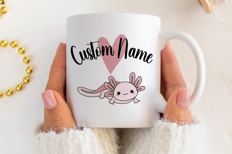 K&ouml;nnte beinhalten: Wei&szlig;e Keramik-Tasse mit einer rosa Axolotl-Illustration und einem rosa Herz. Der Text "Custom Name" ist in schwarzer Schrift geschrieben. Die Tasse hat einen wei&szlig;en Henkel, ideal f&uuml;r Hei&szlig;getr&auml;nke.