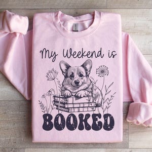 Puede incluir: Una sudadera rosa claro con el texto "My Weekend is Booked" en negro. Un perro corgi de dibujos animados est&aacute; sentado en una pila de libros con flores alrededor.