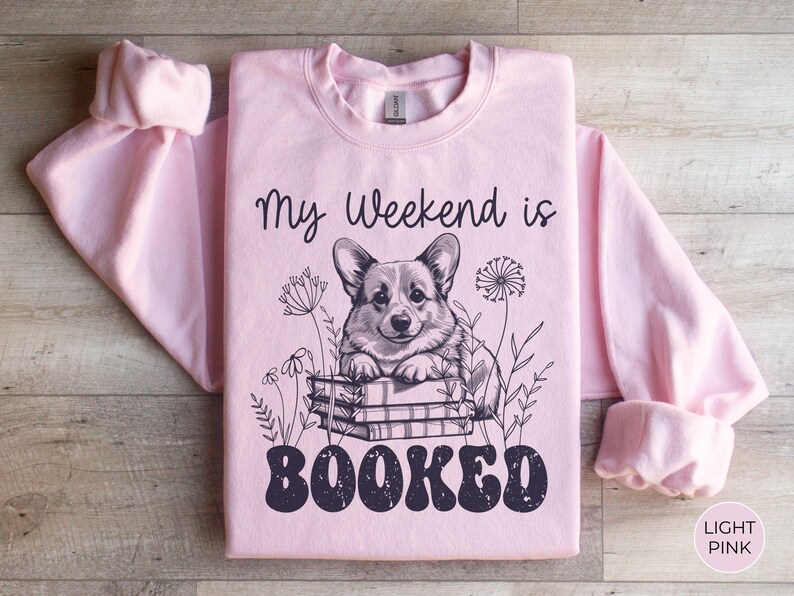 Puede incluir: Una sudadera rosa claro con el texto "My Weekend is Booked" en negro. Un perro corgi de dibujos animados est&aacute; sentado en una pila de libros con flores alrededor.