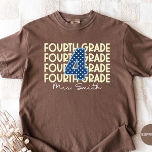 May include: A brown short-sleeve t-shirt featuring the text FOURTH GRADE repeated four times in light yellow. A large dark blue number 4 with white polka dots is centered over the text. The name Mrs. Smith is printed in white cursive below.