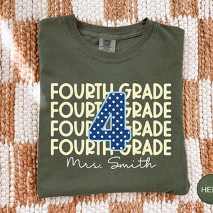 May include: An olive green crew neck t-shirt featuring the repeating text FOURTH GRADE in a light yellow font. A large dark blue number 4 with white polka dots is centered over the text. The name Mrs. Smith is printed in white script below.