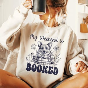 Puede incluir: Una sudadera blanca con un gr&aacute;fico de un perro corgi sentado en una pila de libros. El texto "My Weekend is Booked" est&aacute; impreso en la sudadera.