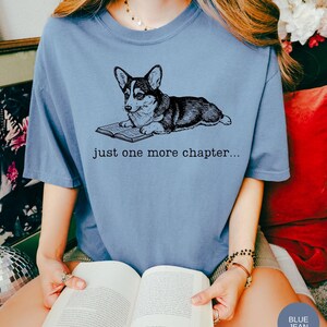 Op de afbeelding: Lichtblauw T-shirt met een zwart-witte illustratie van een Corgi-hond die een boek leest. De tekst "just one more chapter..." staat onder de hond. De persoon draagt een boek.