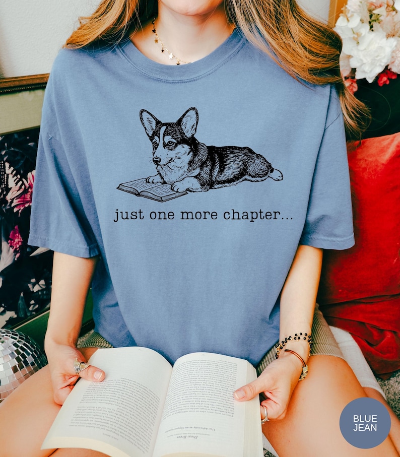 Op de afbeelding: Lichtblauw T-shirt met een zwart-witte illustratie van een Corgi-hond die een boek leest. De tekst "just one more chapter..." staat onder de hond. De persoon draagt een boek.