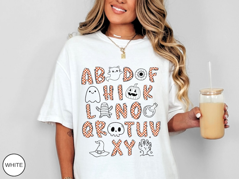May include: A white t-shirt featuring a Halloween-themed alphabet graphic. The letters are filled with an orange and white checkered pattern, interspersed with illustrations of a ghost, pumpkin, eyeball, mummy, potion bottle, skull, hat, and hand.