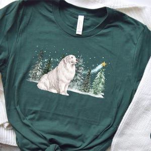 May include: Forest green t-shirt featuring a white Great Pyrenees dog sitting in a snowy forest scene with Christmas trees and a star. The shirt has a crew neck and short sleeves. The word "FOREST" is printed on a green circle.