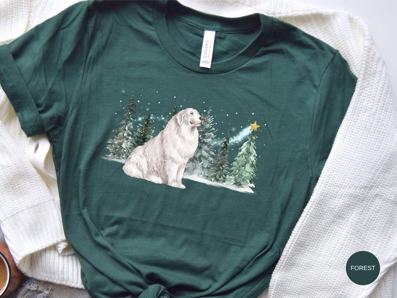 May include: Forest green t-shirt featuring a white Great Pyrenees dog sitting in a snowy forest scene with Christmas trees and a star. The shirt has a crew neck and short sleeves. The word "FOREST" is printed on a green circle.