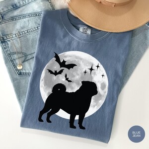 K&ouml;nnte beinhalten: Blaues Comfort Colors T-Shirt mit einem schwarzen Mops-Design vor einem Vollmond, Flederm&auml;usen und Sternen. Das Shirt wird mit hellblauen Jeans und einem beigen Hut pr&auml;sentiert. Die Worte "Blue Jean" sind auf einem blauen Kreis aufgedruckt.