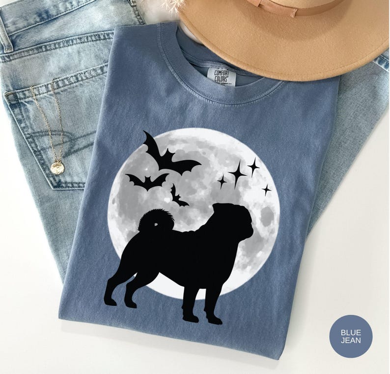 K&ouml;nnte beinhalten: Blaues Comfort Colors T-Shirt mit einem schwarzen Mops-Design vor einem Vollmond, Flederm&auml;usen und Sternen. Das Shirt wird mit hellblauen Jeans und einem beigen Hut pr&auml;sentiert. Die Worte "Blue Jean" sind auf einem blauen Kreis aufgedruckt.