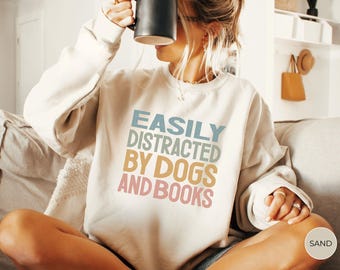 May include: A white sweatshirt with the text "EASILY DISTRACTED BY DOGS AND BOOKS" in colorful letters. The text is in a retro style font.