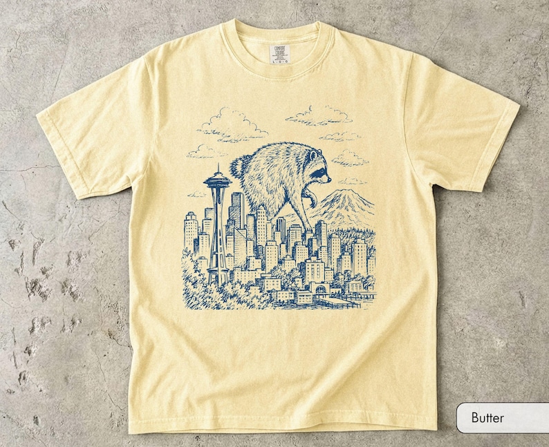 Pu&ograve; includere: Una t-shirt a maniche corte color burro con un'illustrazione grafica blu scuro di un procione gigante che sovrasta lo skyline di Seattle, inclusi lo Space Needle, una montagna sullo sfondo e nuvole sopra gli edifici della citt&agrave;.