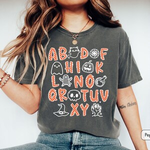 May include: A charcoal gray short-sleeve t-shirt featuring a graphic print of an alphabet where letters are replaced by Halloween icons including a ghost, pumpkin, mummy, skull, witch hat, and potion bottle, all in an orange checkered pattern.