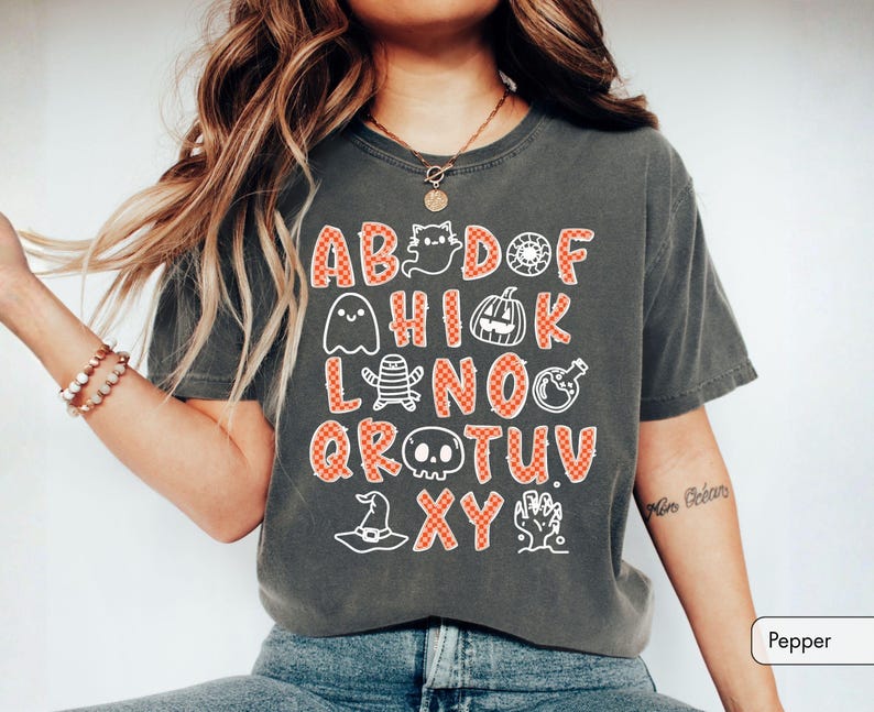 May include: A charcoal gray short-sleeve t-shirt featuring a graphic print of an alphabet where letters are replaced by Halloween icons including a ghost, pumpkin, mummy, skull, witch hat, and potion bottle, all in an orange checkered pattern.