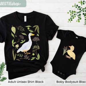 May include: A black adult unisex t-shirt and a black baby bodysuit with a white duck and a yellow duckling illustration. The adult shirt features a white duck with green foliage and flowers. The baby bodysuit features a yellow duckling with green reeds.