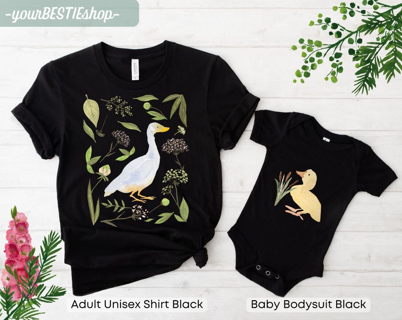 May include: A black adult unisex t-shirt and a black baby bodysuit with a white duck and a yellow duckling illustration. The adult shirt features a white duck with green foliage and flowers. The baby bodysuit features a yellow duckling with green reeds.