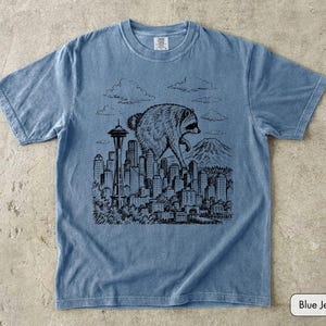Pu&ograve; includere: Una t-shirt a maniche corte color blu jeans con una stampa grafica nera in stile inchiostro che raffigura un procione gigante che sovrasta lo skyline di una citt&agrave;, inclusi lo Space Needle e una montagna sullo sfondo, con nuvole.