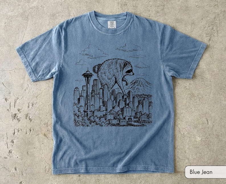 Pu&ograve; includere: Una t-shirt a maniche corte color blu jeans con una stampa grafica nera in stile inchiostro che raffigura un procione gigante che sovrasta lo skyline di una citt&agrave;, inclusi lo Space Needle e una montagna sullo sfondo, con nuvole.