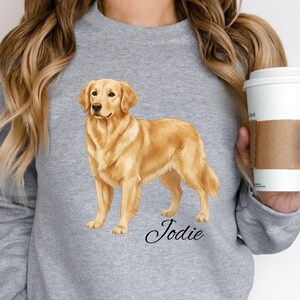May include: Sport grey sweatshirt featuring a golden retriever illustration. The dog is a light brown color and is standing. The name "Jodie" is written in cursive below the dog. A white coffee cup with a brown sleeve is held in the hand.