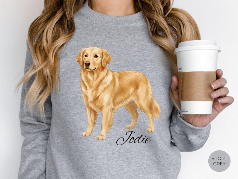 May include: Sport grey sweatshirt featuring a golden retriever illustration. The dog is a light brown color and is standing. The name "Jodie" is written in cursive below the dog. A white coffee cup with a brown sleeve is held in the hand.
