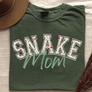 May include: Dark green t-shirt with the text "SNAKE Mom" in a floral design. The word "SNAKE" is in a white, block letter style with a floral pattern. The word "Mom" is in a script font. A brown hat and dried plant are in the background.