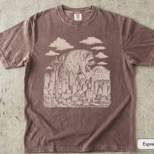 Pu&ograve; includere: Una t-shirt a maniche corte color espresso con una stampa grafica chiara di un procione gigante che sovrasta lo skyline di una citt&agrave; che include lo Space Needle e una montagna, con nuvole sopra e il testo Espresso nell'angolo.