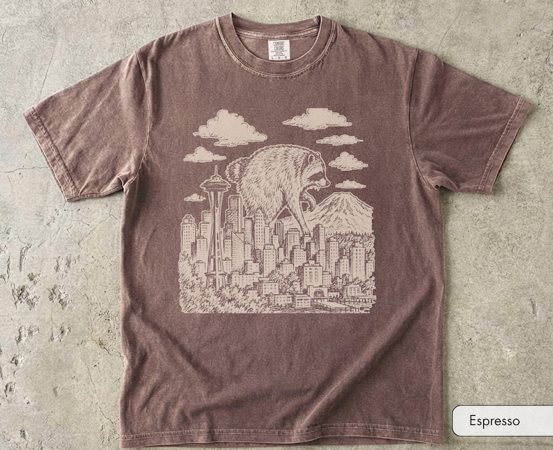 Pu&ograve; includere: Una t-shirt a maniche corte color espresso con una stampa grafica chiara di un procione gigante che sovrasta lo skyline di una citt&agrave; che include lo Space Needle e una montagna, con nuvole sopra e il testo Espresso nell'angolo.