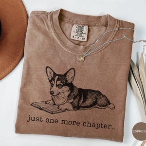 Op de afbeelding: Espresso-kleurige t-shirt met een zwart-wit illustratie van een corgi die een boek leest. De tekst "just one more chapter..." staat eronder. Het shirt heeft een ronde hals en een klein label.