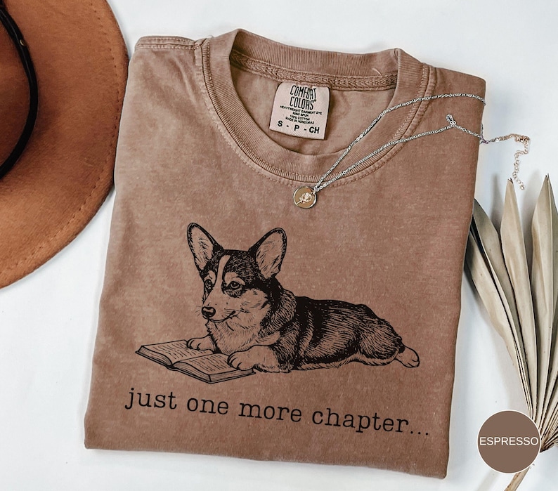 Op de afbeelding: Espresso-kleurige t-shirt met een zwart-wit illustratie van een corgi die een boek leest. De tekst "just one more chapter..." staat eronder. Het shirt heeft een ronde hals en een klein label.