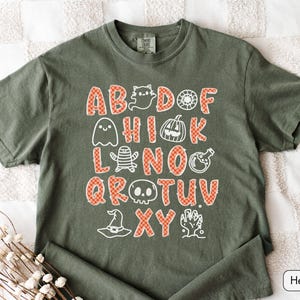 May include: An olive green short-sleeve t-shirt featuring a graphic print of the alphabet with letters in an orange and white checkered pattern. The design includes white line-art illustrations of ghosts, a pumpkin, a mummy, a skull, and a hat.