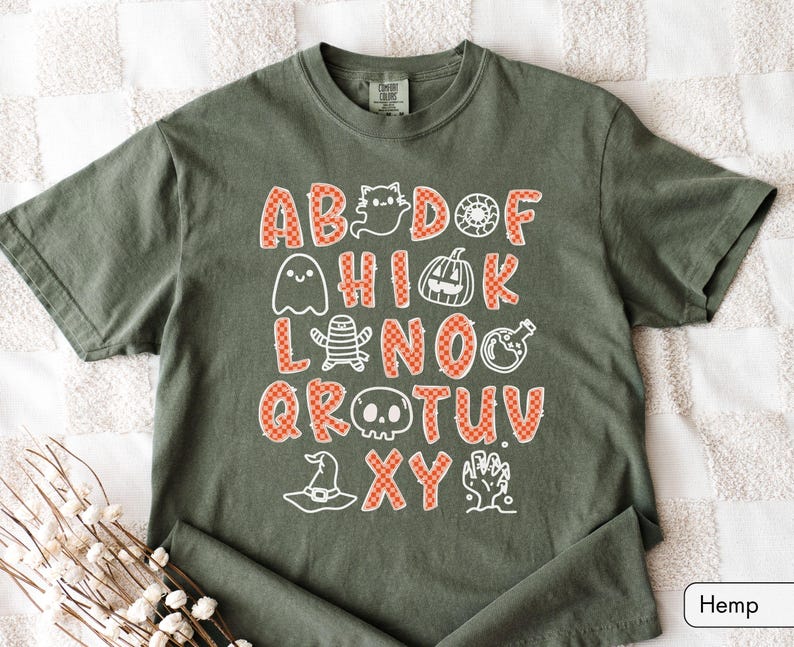 May include: An olive green short-sleeve t-shirt featuring a graphic print of the alphabet with letters in an orange and white checkered pattern. The design includes white line-art illustrations of ghosts, a pumpkin, a mummy, a skull, and a hat.