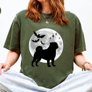 K&ouml;nnte beinhalten: Olivgr&uuml;nes T-Shirt mit schwarzer Silhouette eines Mops vor Vollmond, Flederm&auml;usen und Sternen. Das Shirt hat einen Rundhalsausschnitt und kurze &Auml;rmel. Das Wort "HEMP" ist oben links auf dem Shirt aufgedruckt.