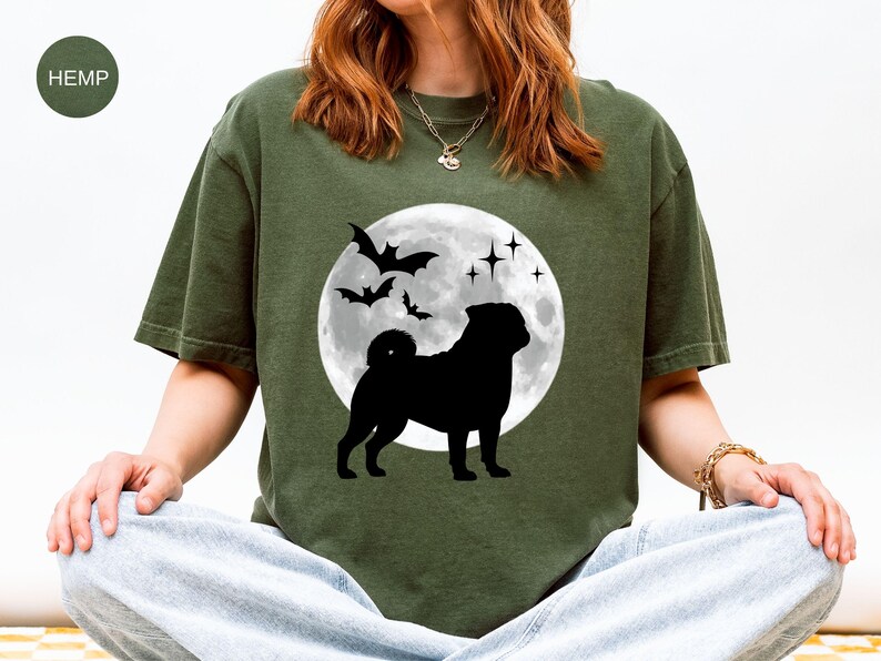 K&ouml;nnte beinhalten: Olivgr&uuml;nes T-Shirt mit schwarzer Silhouette eines Mops vor Vollmond, Flederm&auml;usen und Sternen. Das Shirt hat einen Rundhalsausschnitt und kurze &Auml;rmel. Das Wort "HEMP" ist oben links auf dem Shirt aufgedruckt.