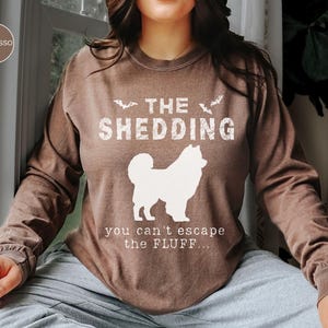 May include: Espresso-colored long-sleeve shirt with the text "THE SHEDDING" and a white dog silhouette. Below the dog, the text reads "you can't escape the FLUFF...". The shirt is a casual wear item.