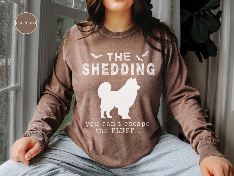 May include: Espresso-colored long-sleeve shirt with the text "THE SHEDDING" and a white dog silhouette. Below the dog, the text reads "you can't escape the FLUFF...". The shirt is a casual wear item.