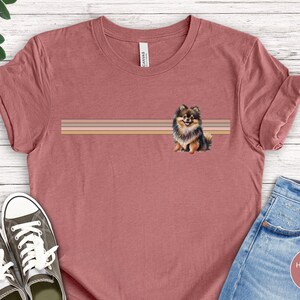 Puede incluir: Una camiseta de color malva jaspeado con una ilustraci&oacute;n de un perro pomerania marr&oacute;n, negro y blanco. El perro est&aacute; de pie sobre un fondo a rayas con tres rayas horizontales en tonos marrones, rosas y beige.