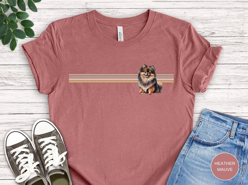 Puede incluir: Una camiseta de color malva jaspeado con una ilustraci&oacute;n de un perro pomerania marr&oacute;n, negro y blanco. El perro est&aacute; de pie sobre un fondo a rayas con tres rayas horizontales en tonos marrones, rosas y beige.