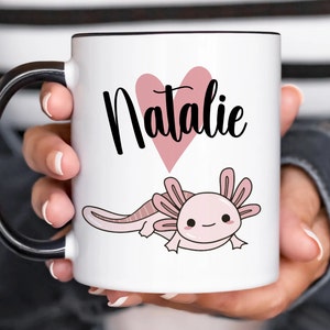 K&ouml;nnte beinhalten: Wei&szlig;er Keramikbecher mit schwarzem Henkel und Rand. Der Becher zeigt den Namen "Natalie" in schwarzer Schrift &uuml;ber einem rosa Herz und eine Cartoon-Axolotl-Illustration in Rosa.